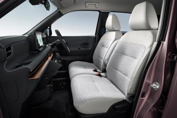 「日産サクラ」の車内を、後部ドア側から前方に向けて斜めに撮影した画像。明るいグレーの前席・後席シートが並び、前方には黒いダッシュボードとハンドルが見える。背景は白で、室内空間全体が確認できる。