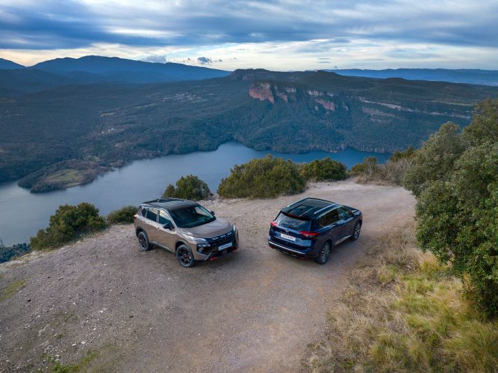 Zwei neue Nissan X‑Trail SUVs des Modelljahres 2026 stehen auf einer Lichtung mit Blick auf einen großen See, umgeben von bewaldeten Hügeln und Bergen unter einem bewölkten Himmel. Die Fahrzeuge, ein N-Trek in der Karosseriefarbe Baja Storm und ein Tekna in der Karosseriefarbe Universal Blue, sind so positioniert, dass sie von vorne und hinten zu sehen sind und ihr Design und ihre Geländegängigkeit in einer malerischen Außenkulisse zur Geltung bringen.