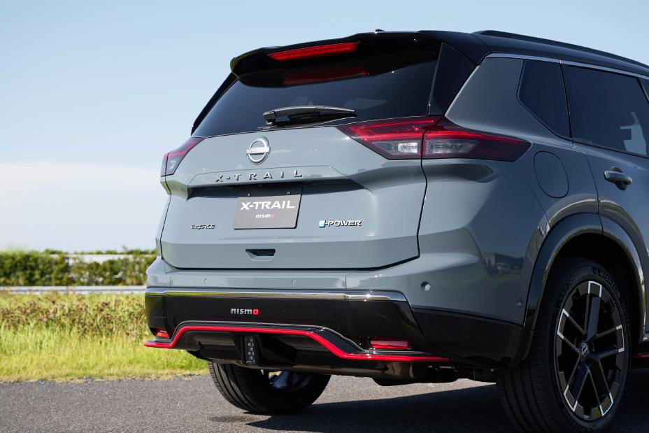 A grey Nissan X-Trail NISMO is parked on a road. The vehicle is captured from the rear, featuring black and red accents including red trim on the lower bumper and around the exhaust. The background includes greenery and an open sky.