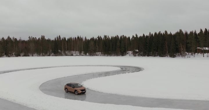 Elektroauto Nissan Ariya e‑4ORCE in Akatsuki Copper mit Dach und Außenspiegel in Pearl Black fährt auf Eissee mit schneebedeckten Bäumen im Hintergrund, 3/4 Heck, Drohnen-Perspektive