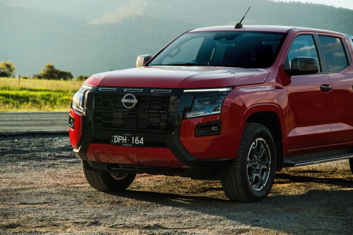 Front three-quarter close view of a 2026 Nissan Navara ST‑X dual cab painted in Outback Red, parked on gravel with the black grille, front wheel, and cabin side visible.