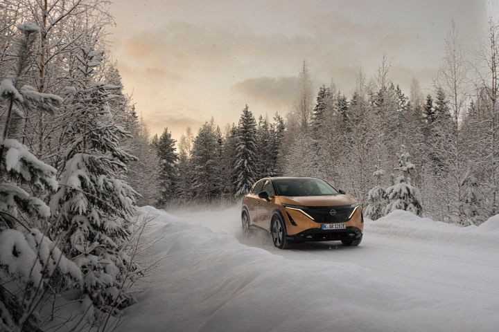 Elektroauto Nissan Ariya e‑4ORCE in Akatsuki Copper mit Dach und Außenspiegel in Pearl Black fährt auf schneebedeckter Straße mit Schneehaufen und verschneiten Bäumen auf beiden Seiten, 3/4 Front