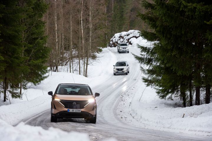 Elektroauto Nissan Ariya e‑4ORCE in Akatsuki Copper mit Dach und Außenspiegel in Pearl Black  fährt auf schneebedeckter Straße vor zwei silbernenNissan X‑Trail e‑Power e‑4ORCE