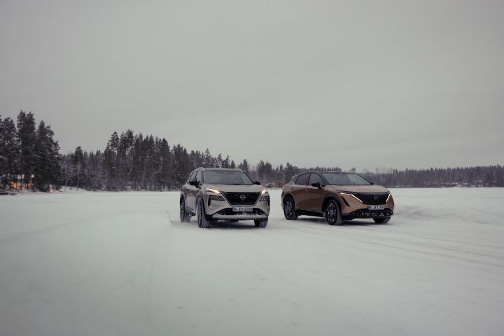 Silberner Nissan X‑Trail e‑Power e‑4ORCE und Elektroauto Nissan Ariya e‑4ORCE in Akatsuki Copper mit Dach und Außenspiegel in Pearl Black fahren nebeneinander auf schneebedecktem EIssee mit verschneitem Wald im Hintergrund, 3/4 Front