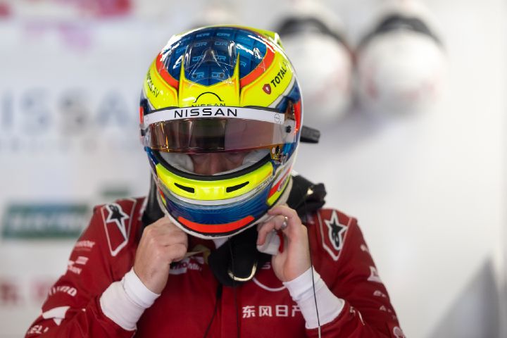 Oliver Rowland setzt seinen gelb-blauen Helm mit hochgeklapptem Visier auf, während er in der Teamgarage seinen roten Rennanzug trägt.