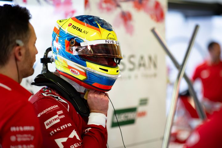 Oliver Rowland setzt seinen Helm auf, während er seinen roten Nissan Formula E Team-Rennanzug in der Teamgarage außerhalb des Autos trägt.