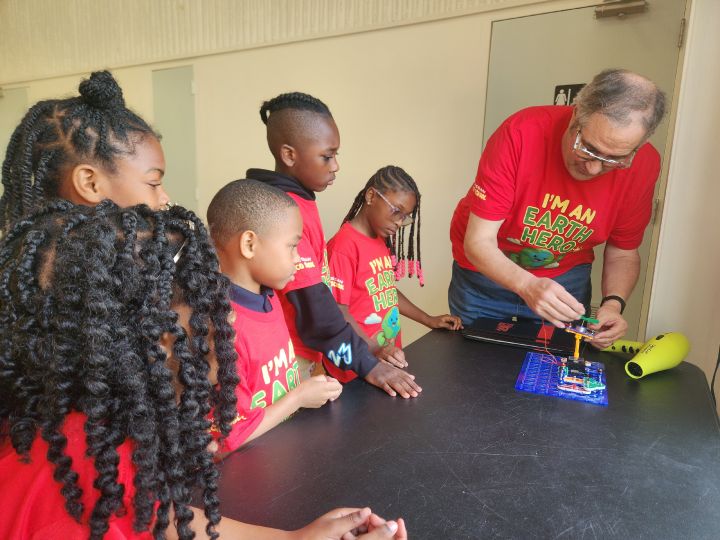 A small group of children stand observing a Nissan employee put together a project.