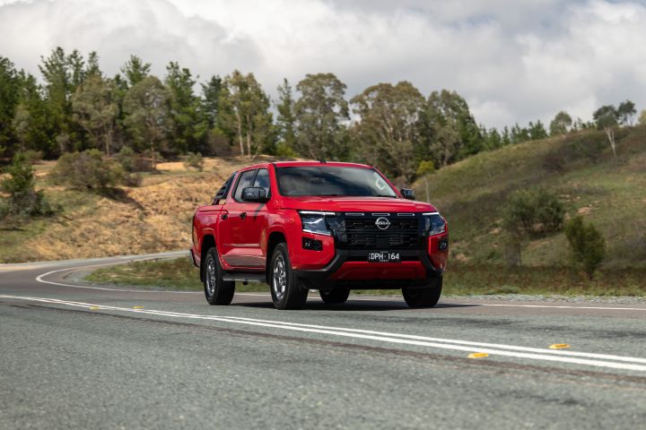 A red 2026 Nissan Navara ST‑X travelling on a multi‑lane road, with hills and scattered trees lining the landscape.