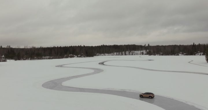 Elektroauto Nissan Ariya e‑4ORCE in Akatsuki Copper mit Dach und Außenspiegel in Pearl Black fährt auf Eissee mit schneebedeckten Bäumen im Hintergrund, kleines Auto in weißer Landschaft aus der Drohnen-Perspektive