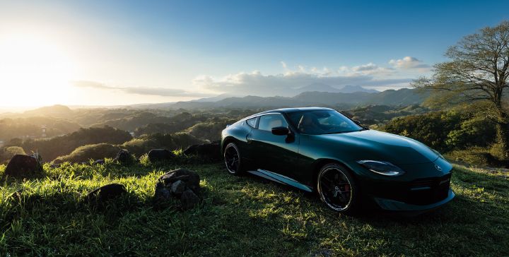 Unryu Green Fairlady Z parked on a grassy hillside, photographed from a front-right three-quarter angle. The vehicle faces toward the right side of the image, showcasing its long-hood, short-deck sports car proportions, black alloy wheels, and red brake calipers. Rolling mountains and forested landscapes stretch across the background, while soft sunlight illuminates the grass and bodywork. Set against a clear blue sky and expansive natural scenery, the rich Uryu Green exterior finish stands out prominently.