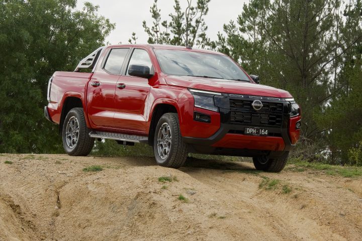 Front three‑quarter driving view of a 2026 Nissan Navara ST‑X Outback Red dual cab travelling on a sealed country road with grassy slopes and trees nearby.
