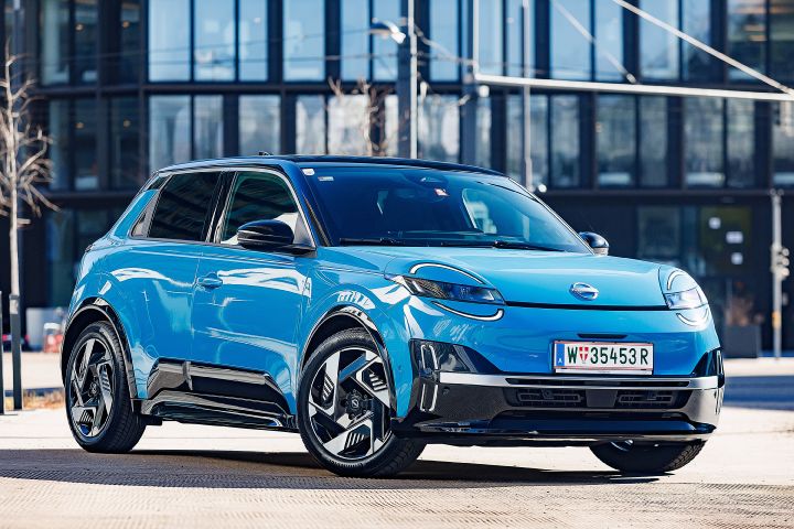 3/4-Frontansicht eines leuchtend blauen Nissan Micra mit schwarzem Kontrastdach, der vor einem Bürogebäude mit gläserner Front parkt