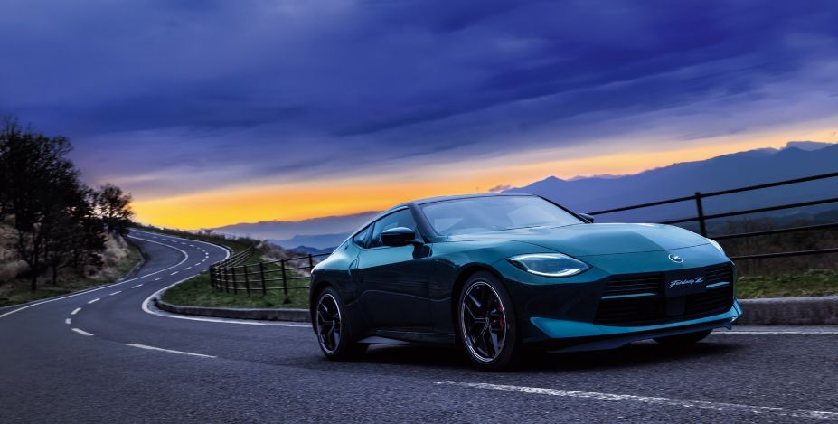 雲龍グリーンのフェアレディZ。ワインディングロード沿いに停車する車両を、右前方からの3/4アングルで撮影している。車両前方は右方向を向き、低く構えたフロントノーズやロングノーズ・ショートデッキのシルエット、ブラックのアルミホイールが際立つ。背景には大きくカーブする道路とガードレール、遠くの山並みが広がり、夕暮れ時の空には濃い雲とオレンジ色の地平線が見える。ドラマチックな空模様の下で、深みのある雲龍グリーンのボディカラーが印象的に映える写真。