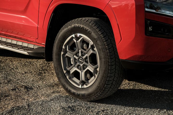 Close-up of the front wheel and lower front guard on a 2026 Nissan Navara ST-X in Outback Red, showing the alloy wheel design, tyre sidewall, black wheel-arch trim, and side step on gravel.