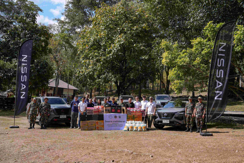 A group of people standing outdoors in front of two Nissan vehicles, surrounded by trees. They are posing with donated supplies arranged on tables and the ground, with two tall Nissan flags on each side.