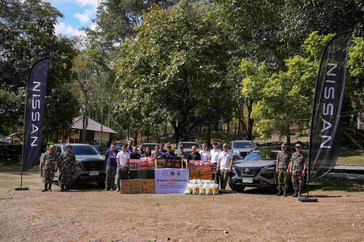 A group of people standing outdoors in front of two Nissan vehicles, surrounded by trees. They are posing with donated supplies arranged on tables and the ground, with two tall Nissan flags on each side.