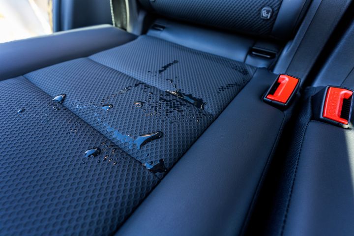 Der neue 2026 Nissan X‑Trail N-Trek zeigt Wassertropfen auf Ledersitzen, die mit wasserdichtem CellCloth®-Bezugstoff bezogen sind, mit sichtbaren magmaroten Sicherheitsgurt-Schnallen. Das Bild verdeutlicht die Wasserbeständigkeit bzw. die Auswirkungen von Verschüttungen auf den Bezugsstoff des Fahrzeugs.