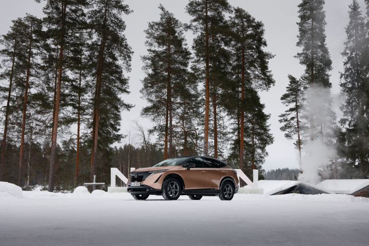 Blick von unten auf das Elektroauto Nissan Ariya e‑4ORCE in Akatsuki Copper mit Dach und Außenspiegel in Pearl Black im Schnee vor weißem Nissan Schriftzug