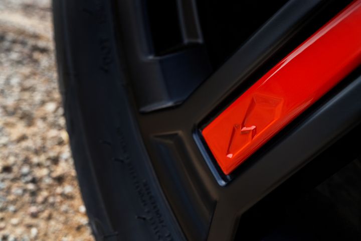 Nahaufnahme des neuen 2026 Nissan X‑Trail Tekna mit schwarzem Reifen und Felge mit magmarotem reflektierendem Detail und kleinem, geprägtem Mount-Fuji-Symbol. Das Bild hebt die Kontraste zwischen der rauen Bodenoberfläche, dem glatten Reifengummi und dem glänzenden roten Akzent auf der Felge hervor.