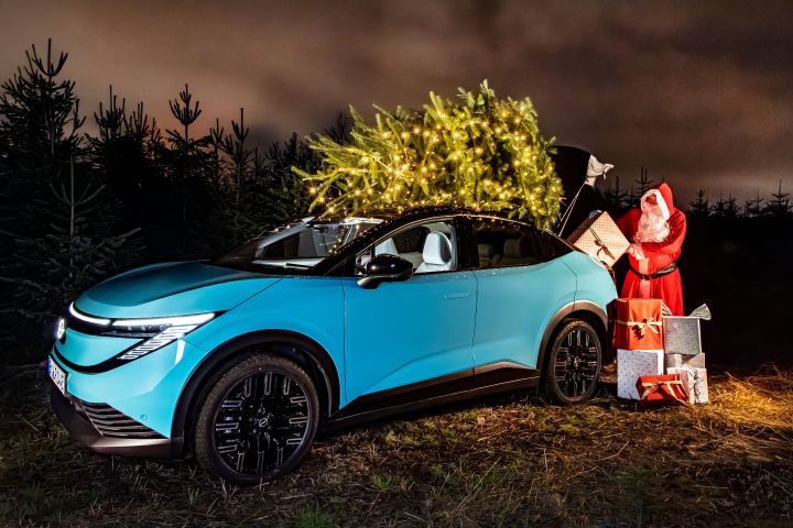 Ein türkisblauer Nissan LEAF steht vor einer Weihnachtsbaumplantage, auf dem Dach ein beleuchteter Weihnachtsbaum, der Weihnachtsmann lädt Geschenke in den Kofferraum