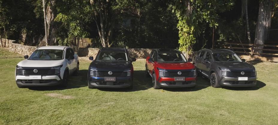 Quatro carros Nissan Kicks, nas cores branca, preta, vermelha e cinza escuro, estão estacionados lado a lado em uma área gramada com árvores ao fundo. Os veículos exibem o logotipo da Nissan na grade frontal