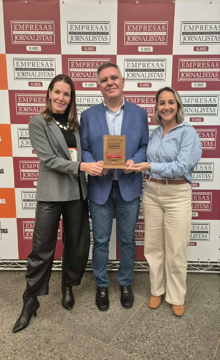 Três pessoas da equipe de Comunicação Corporativa da Nissan do Brasil seguram um troféu retangular à frente de um fundo quadriculado com a marca da Negócios da Comunicação