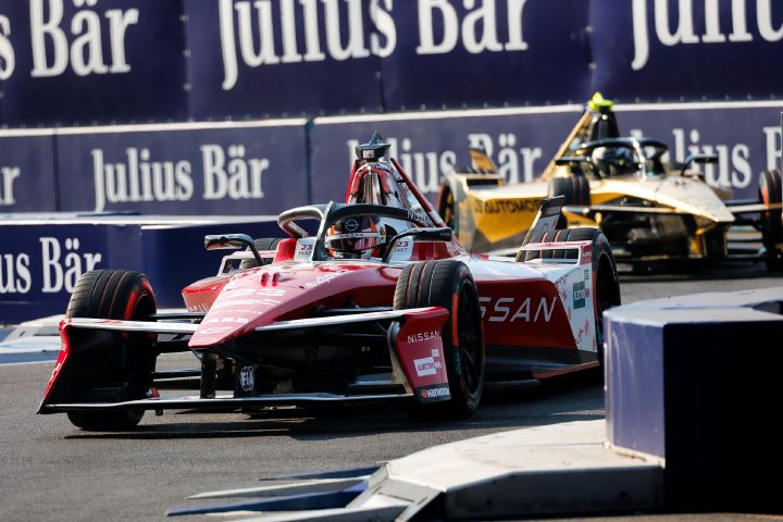 Ein rot-weißer Nissan Formel-E-Rennwagen fährt auf einer Rennstrecke nach links in den Vordergrund. Im Cockpit ist Norman Nato zu sehen, hinter ihm folgt ein weiterer Formel-E-Rennwagen. Im Hintergrund sind Sicherheitsbarrieren und Sponsorenlogos zu erkennen.