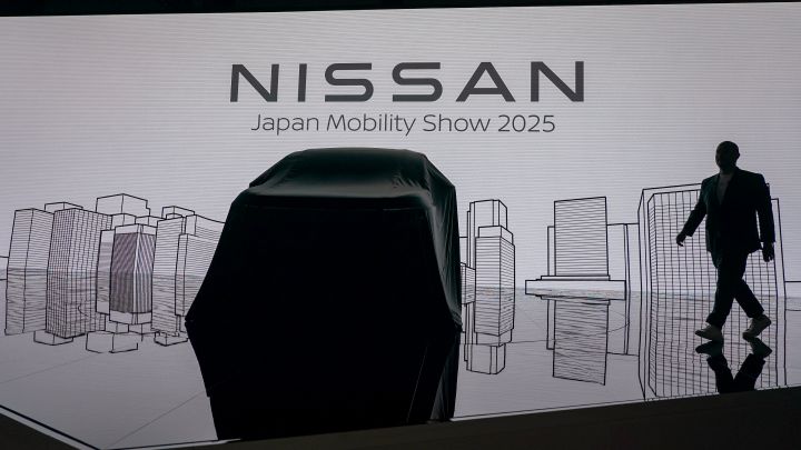 In a dark stage setting, a vehicle covered with a black cloth is positioned at the center, while the background screen displays the text “Nissan Japan Mobility Show 2025” along with cityscape line art. On the right side, CEO Ivan Espinosa in a suit is walking.