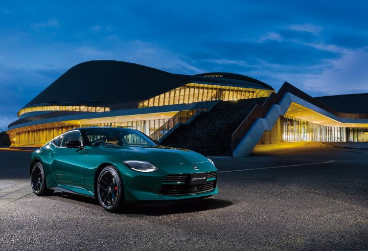Unryu Green Fairlady Z parked in front of a large illuminated modern building at dusk, photographed from a front-right three-quarter angle. The vehicle faces toward the right side of the image, showcasing its low-slung front end, long-hood, short-deck proportions, black alloy wheels, and red brake calipers. In the background, a contemporary architectural structure with a sweeping dark roof and expansive glass walls glows with warm interior lighting. The contrast between the illuminated building and the deep blue evening sky highlights the rich Unryu Green exterior finish and the distinctive styling of the Fairlady Z.