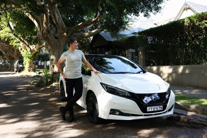Nissan Australia ambassador Osher Gunsberg stands next to his white Nissan LEAF and looks to his left while standing on the street