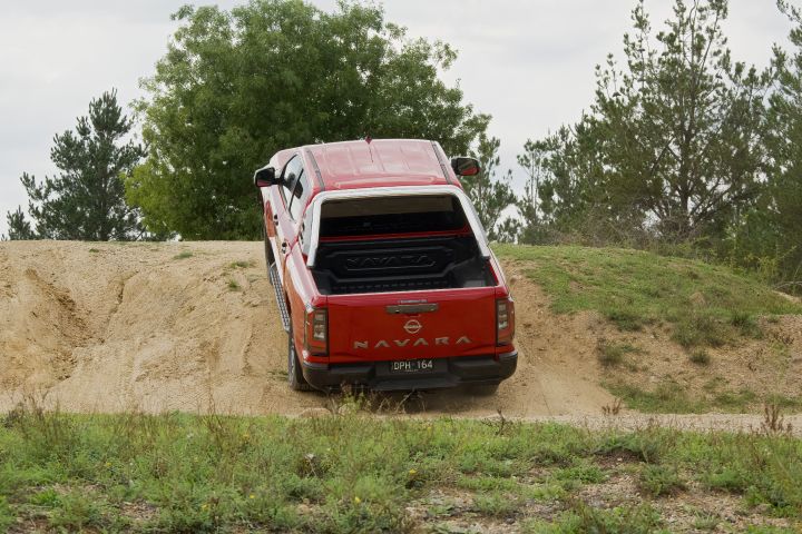 Rear view of a 2026 Nissan Navara ST‑X dual cab finished in Outback Red, driving up a sandy off‑road rise surrounded by native vegetation.