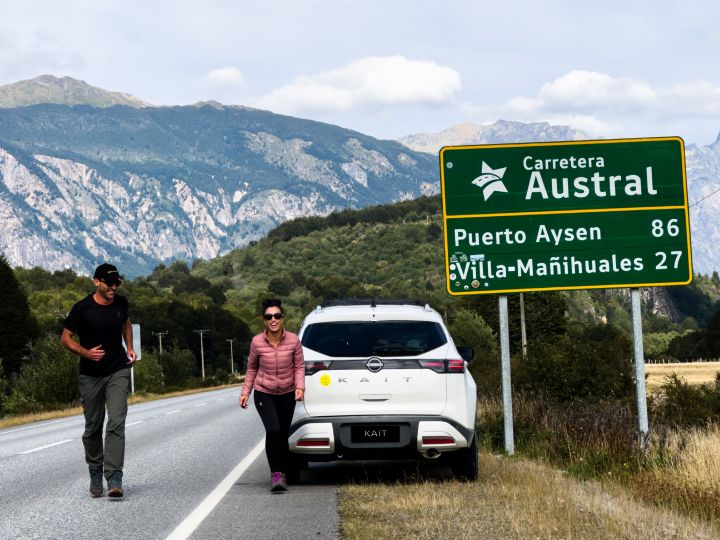 O casal Guilherme e Camila caminham ao lado do Nissan Kait estacionado no acostamento de uma rodovia e ao lado de uma placa onde pode-se ler “Carretera Austral, Puerto Aysen e Villa-Mañihuales