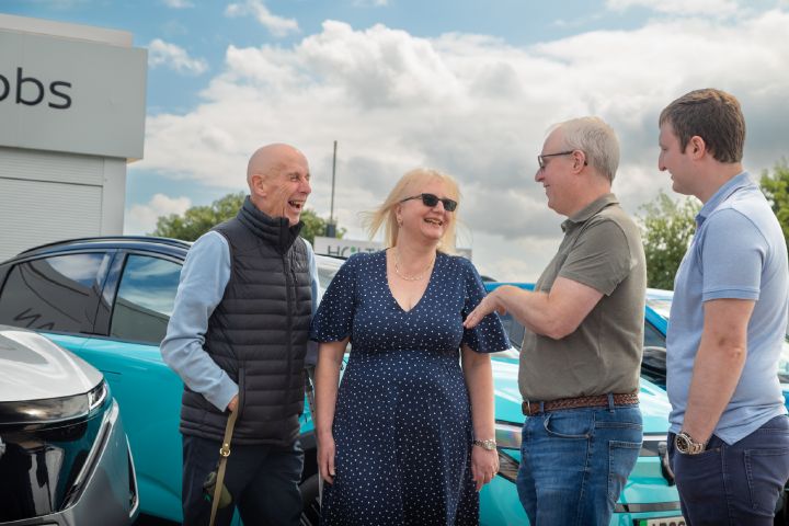 Four people standing and talking beside Nissan vehicles outside a Nissan dealership