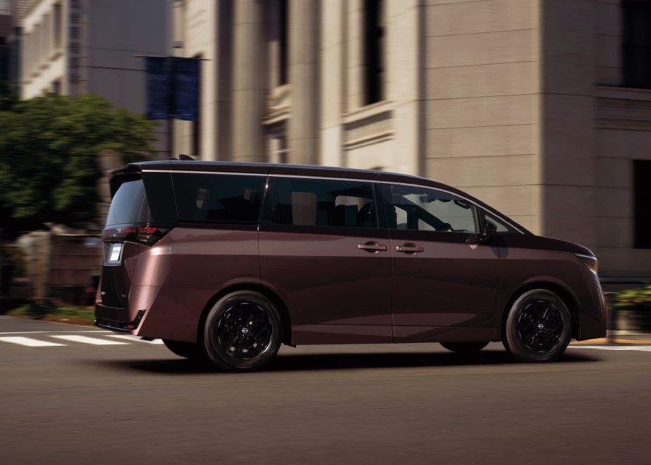A dark brown, slightly burgundy-colored All new ELGRAND driving rightward on a city street. The vehicle is shown mainly from the left side in a panning shot, highlighting its long profile, large windows, and black wheels. Beige high-rise buildings and green trees line the background, with motion blur emphasizing movement. Warm sunlight reflects off the bodywork, showcasing the premium styling and sleek side silhouette of the minivan.
