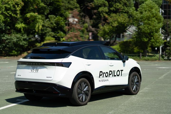 Ein weißer Nissan Ariya wird von der linken Rückseite fotografiert, wobei die Logos „ProPILOT” und „NISSAN” an der Seite zu sehen sind. Das Fahrzeug steht auf einem parkplatzähnlichen Gelände, das von üppig grünen Bäumen und Gebäuden umgeben ist.