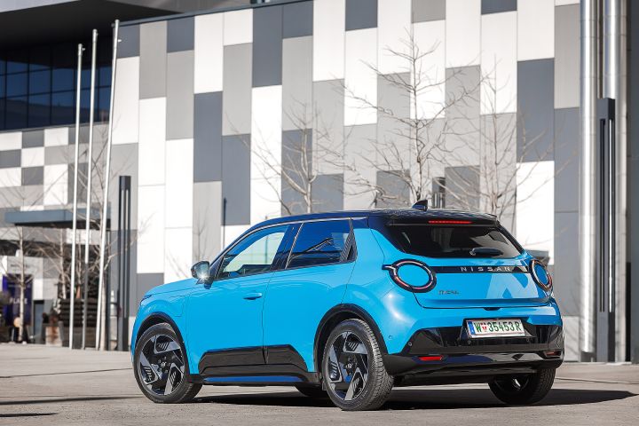 Blick von schräg hinten auf einen parkenden, leuchtend blauen Nissan Micra mit schwarzem Kontrastdach vor einem Gebäude mit grau-weißer Fassade