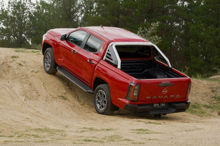 Rear three‑quarter view of a red 2026 Nissan Navara ST‑X driving up a sandy incline in an off‑road environment.