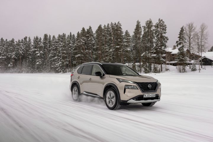 Silberner Nissan X‑Trail e‑Power e‑4ORcE fährt auf schneebedecktem Eissee mit verschneiten Bäumen und Holzhütten im Hintergrund, 3/4 Front