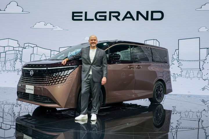 On the stage, the all-new Elgrand in Fuji Dawn and Shigoku two-tone colors is positioned diagonally, facing slightly to the left. Next to the vehicle stands CEO Ivan Espinosa in a suit, resting one hand on the car while posing. The background features a white screen with cityscape line art and the text ELGRAND.