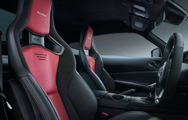 Interior view of a Fairlady Z NISMO, photographed from the rear passenger-side area looking forward. Prominently featured are the NISMO-exclusive black and red RECARO sport seats, with the NISMO logo embroidered on the headrests and RECARO branding integrated into the seatbacks with large harness openings. The right-hand-drive steering wheel, six-speed manual transmission shifter, and center console are also visible. Red contrast stitching accents the predominantly black cabin, while the neutral studio background highlights the performance-oriented seating and driver-focused cockpit design of the Fairlady Z NISMO.