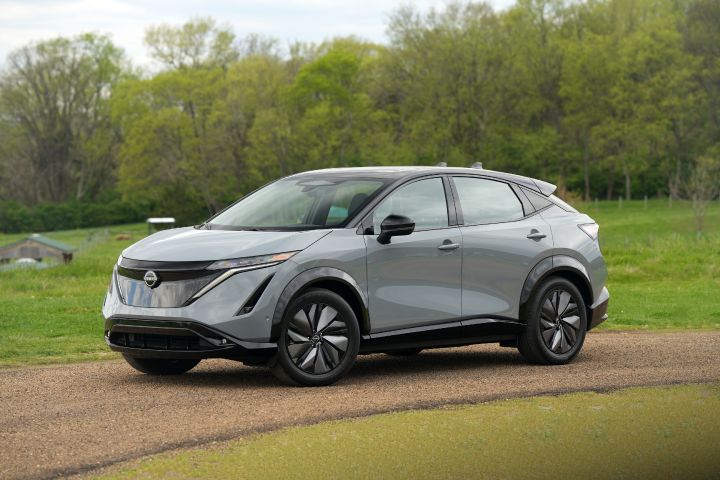 Front left 3/4 view of a gray 2024 Nissan Ariya parked on a dirt road with green pastures in the background.