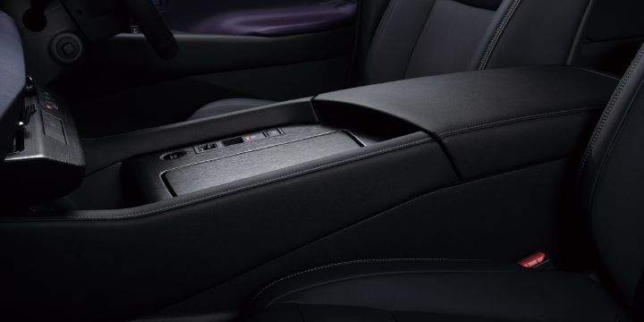 Close-up interior view of the All-new ELGRAND front center console. The premium black-themed cabin features a large center armrest and a wide center console positioned between the front seats. A brushed-metal style sliding cover conceals a storage compartment, while USB ports and control switches are located just above it. Soft-touch materials and stitched trim surfaces enhance the upscale appearance of the console area. Portions of the black leather-style front seats are visible on either side, and the subdued lighting emphasizes the combination of functionality and premium craftsmanship within the cabin.