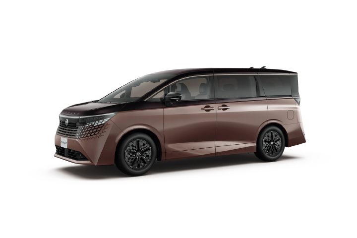 A studio image of the All new ELGRAND in a dark brown metallic finish with a subtle burgundy tone, set against a light gray background. The vehicle is shown facing left from a front-left three-quarter angle, revealing the front fascia and left side profile. A black roof and dark window glass create a two-tone appearance, while the large grille and sleek headlamp design emphasize a modern, sophisticated look. The long body, sculpted side surfaces, and distinctive geometric alloy wheels highlight the premium character and strong presence of the minivan.