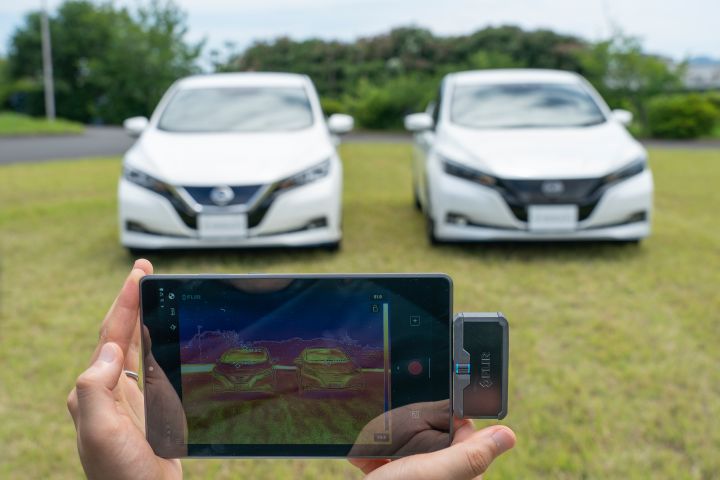 Two white Nissan LEAF models parked on grass with a person's hands in the foreground holding a device showing the temperatures on the cars' surface