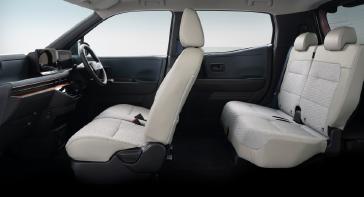 「日産サクラ」の車内を、後席側から前方に向けて撮影した画像。明るいグレーの前席と後席シートが見え、前方にはハンドルとインパネが配置されている。背景は白く、室内レイアウトが分かる構成となっている。