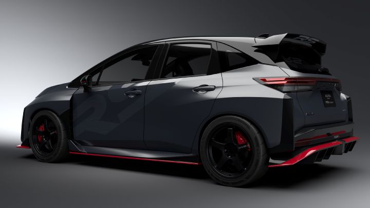 A silver and black two-tone sports car, the Aura Nismo RS Concept, photographed in a studio from a rear-left angled view. The vehicle is stationary, featuring a black plate with white text reading 'AURA NISMO RS CONCEPT' at the center of the rear, along with a red accent line and the NISMO logo on the lower bumper. A large rear wing is mounted, and the side displays a light gray  'RS.' The background is a simple gray gradient, highlighting the car's sporty design