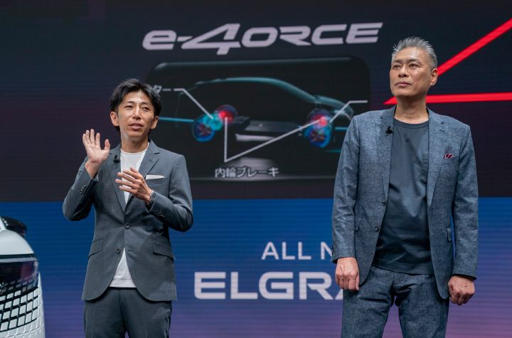 発表会場のステージに立つ中村CPS（左）と一野CVE（右）。中村CPSはグレーのジャケットと白いシャツを着用し、正面やや右を向いて片手を上げながら説明している。一野CVEは濃いグレーのジャケットとTシャツ姿で正面を向いて立っている。背景の大型スクリーンには「e‑4ORCE」のロゴと、新型エルグランドの駆動システムを示すイラストが表示されている。画面下部には「ALL NEW ELGRAND」の文字が見え、左端にはホワイトの新型エルグランドのフロント部分が写っている。青と紫を基調とした照明がステージ全体を照らし、新型エルグランドの技術プレゼンテーションの様子を捉えた写真。