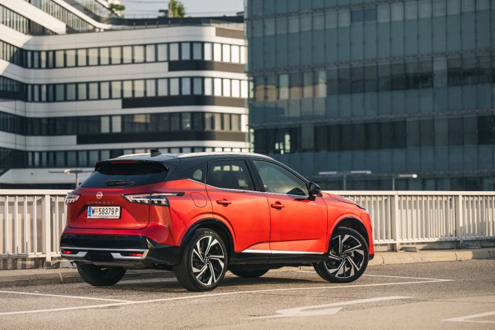 Blick von schräg hinten auf einen parkenden Nissan Qashqai in Fuji Sunset Red vor Hochhäusern
