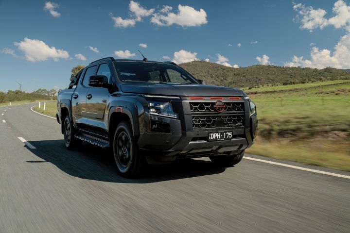 2026 Nissan Navara PRO-4X finished in Boulder Grey shown in a front three-quarter driving shot with black wheels, side steps and red-highlighted grille trim.