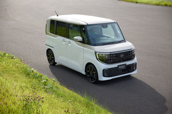 The new Nissan Roox kei car, in white with black wheels and front grille, is driving on a paved road that curves slightly to the left. The road is bordered by green grass and small plants, and the vehicle is captured from an elevated angle.